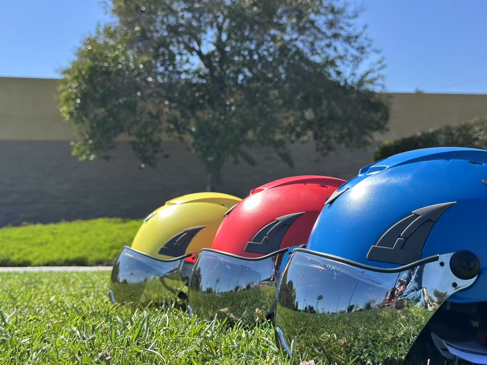Low angle side view of three AeroPro Safety Helmets with Smoke Visor Kit, colors yellow, red and blue on Grass. Three ANSI/ISEA Z89.1-2014 Type 1 Class C Safety Helmets with Smoke Visor used for Construction, Rescue & Arborist. 