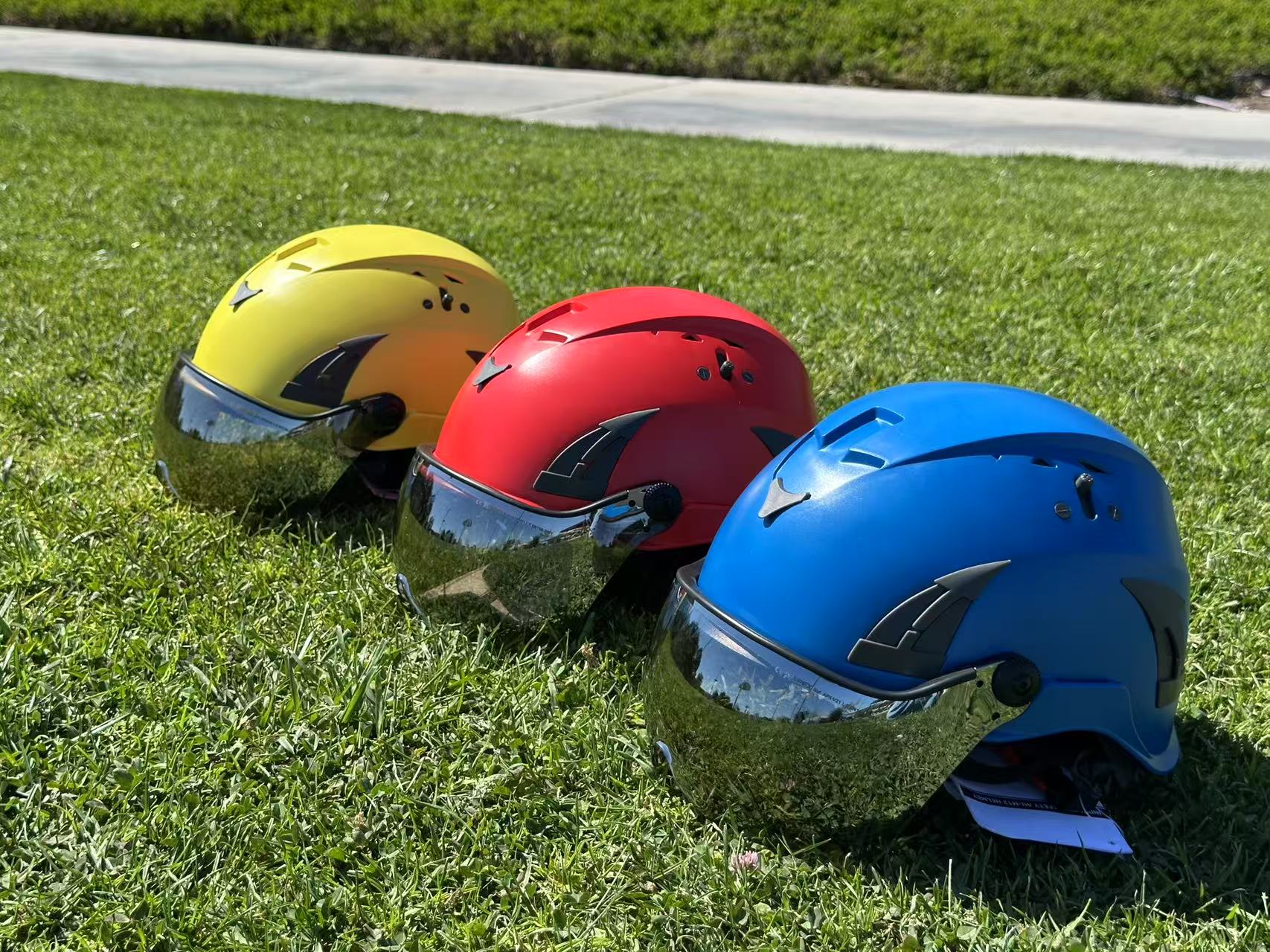 Low angle top down side view of three AeroPro Safety Helmets with Smoke Visor Kit, colors yellow, red and blue on Grass. Three ANSI/ISEA Z89.1-2014 Type 1 Class C Safety Helmets with Smoke Visor used for Construction, Rescue & Arborist. 